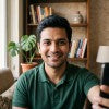 A man wearing a dark green crewneck t-shirt, smiling and looking directly at the camera in a cozy indoor setting with a bookshelf in the background.