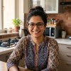 A woman with glasses and a patterned top smiling in a warm, brightly lit kitchen setting.